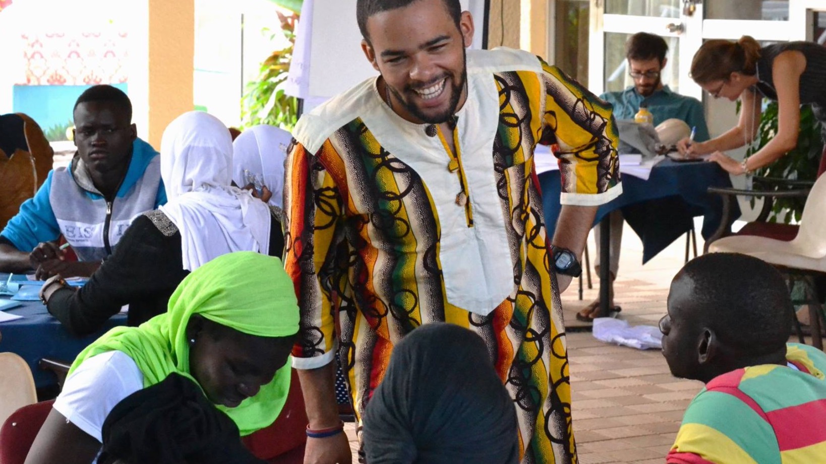 A Peace Corps volunteer smiles while engaging with a group of students at an outdoor classroom setting, as learners participate in a writing activity.