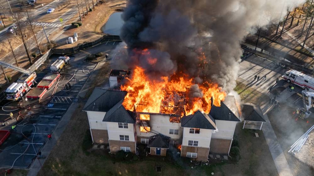 An aerial view of the former UNCP Village Apartments building engulfed in flames during a controlled live-burn demolition exercise, with fire trucks, hoses and first responders visible in the surrounding parking lot.
