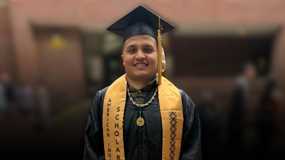 Justice Locklear stands in graduation regalia, wearing a black cap and gown with a gold stole, smiling for a portrait.