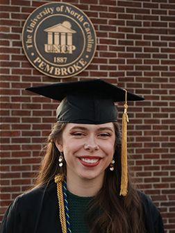 Tiana Lowry smiling at camera in graduation cap in front of a brick wall with the UNCP logo over her right shoulder