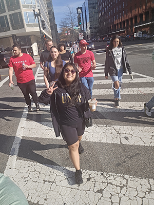 A UNCP student flashes a peace sign while crossing a Washington, D.C. street in a UNCP sweatshirt, with fellow Study Away trip participants visible behind her.