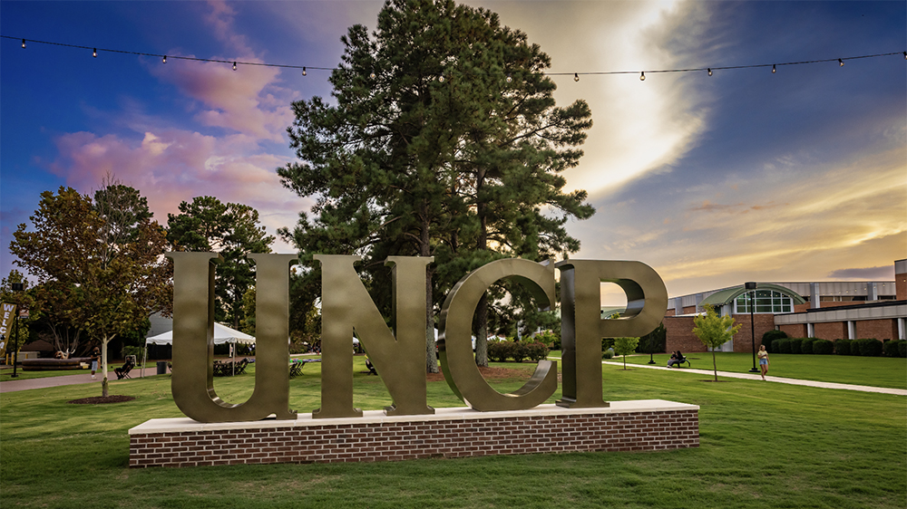 large, golden UNCP letters on the campus of UNCP at sunset with a purple/pink sky