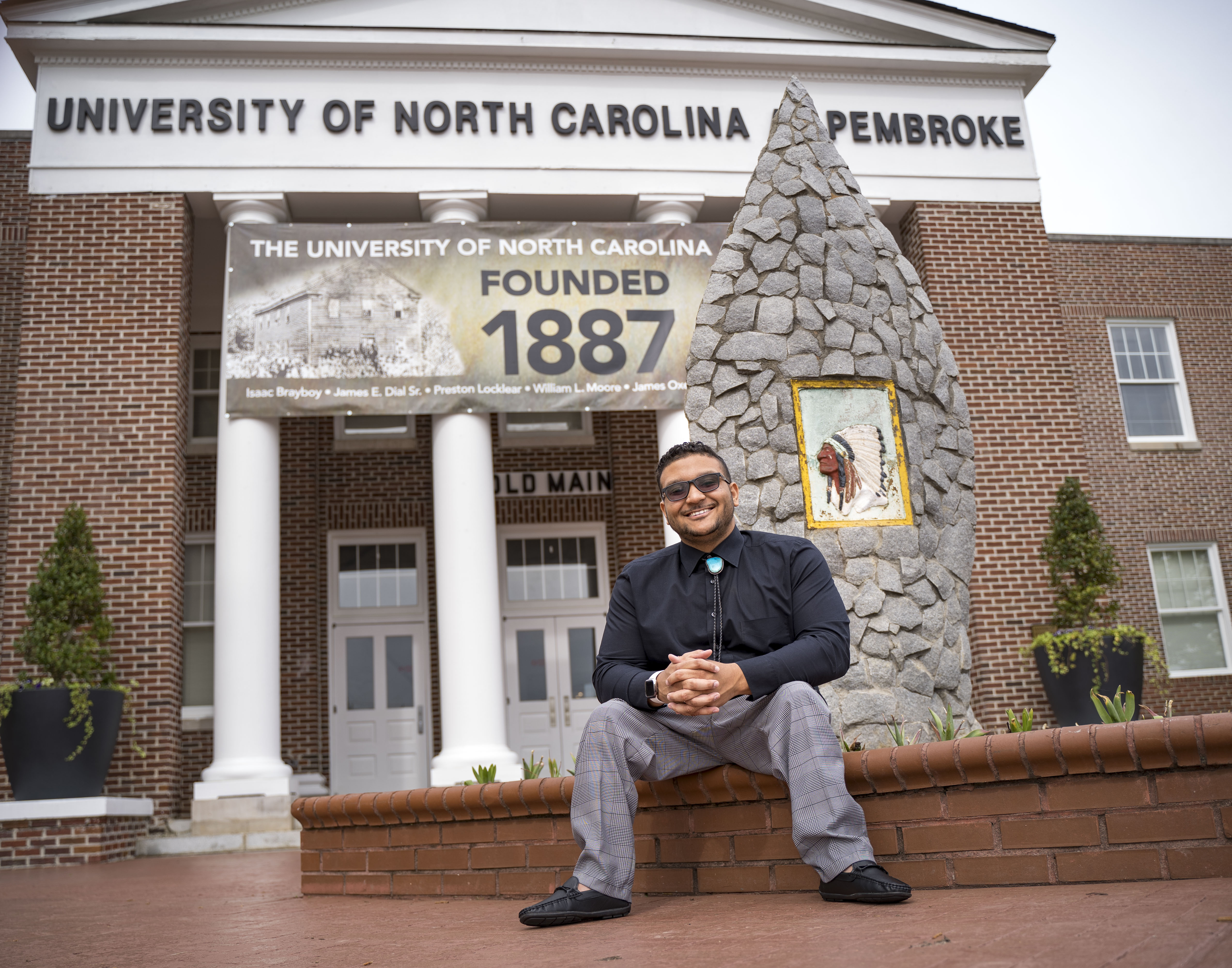 Benjamin Clark sitting and smiling in front of the Old Main building at UNC Pembroke, next to a stone monument with a painted image of a Native American chief.