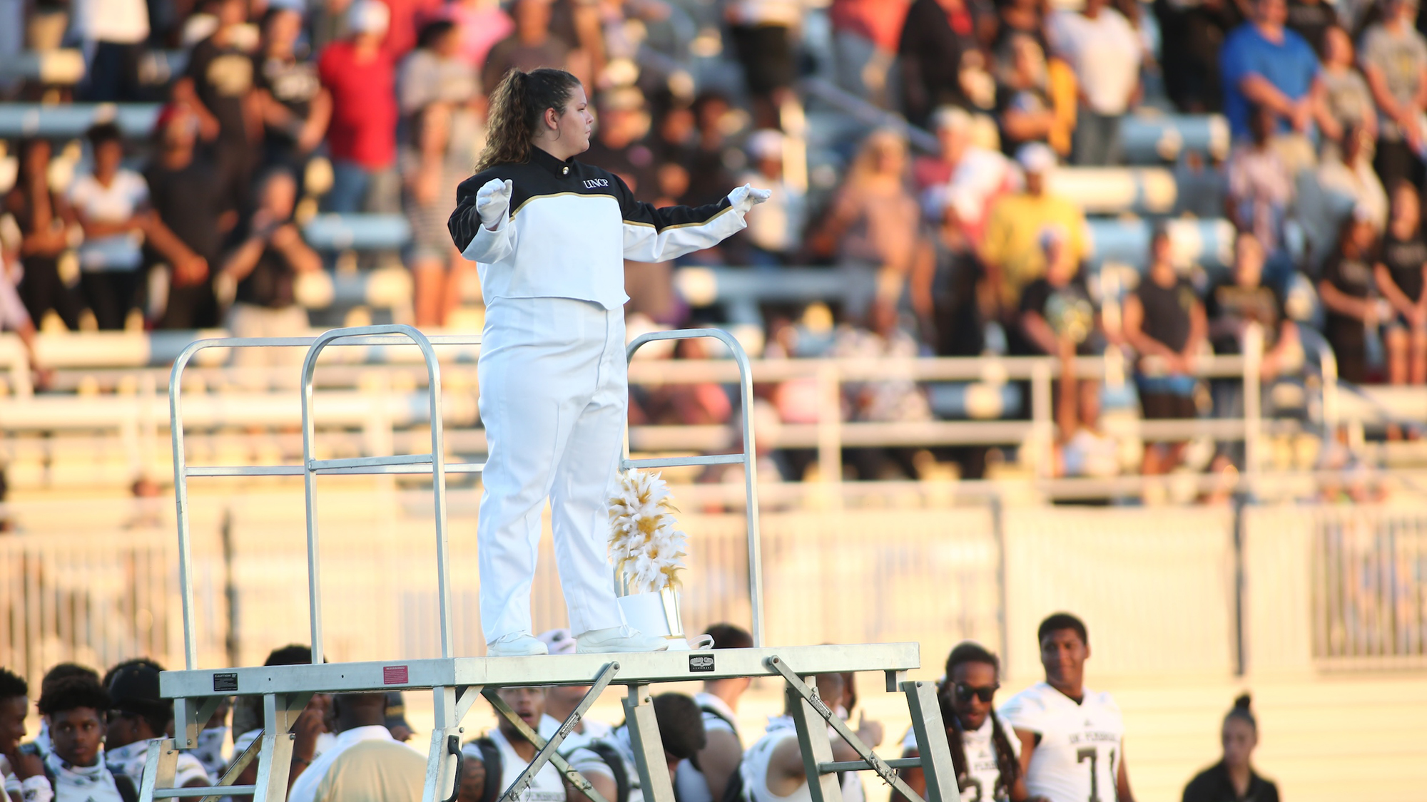 conductor on elevated stand performs at football game with crowd in the stands behind her