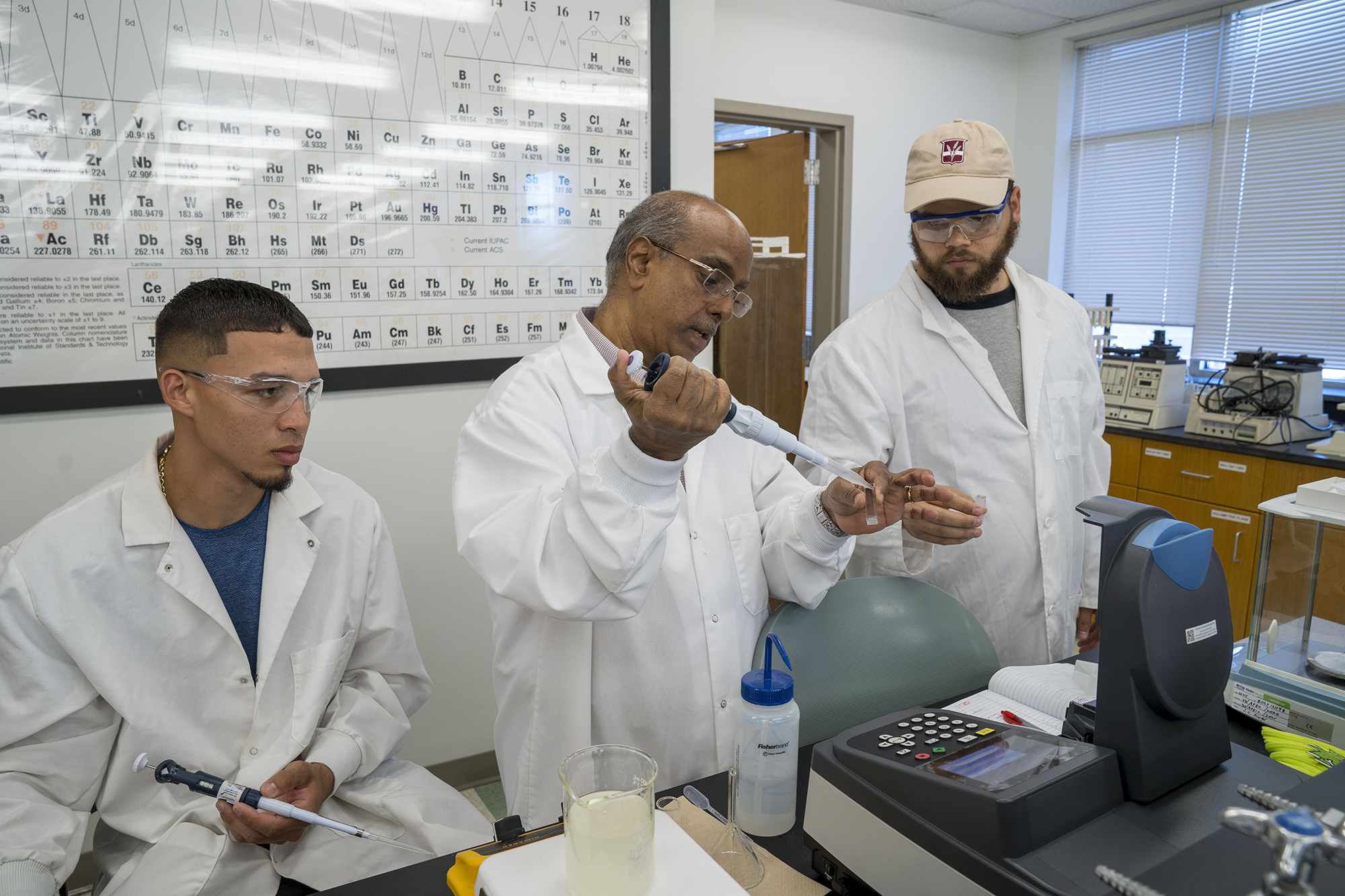 chemistry teacher stands between two male UNCP students in while lab coats showing them how to use a pipette