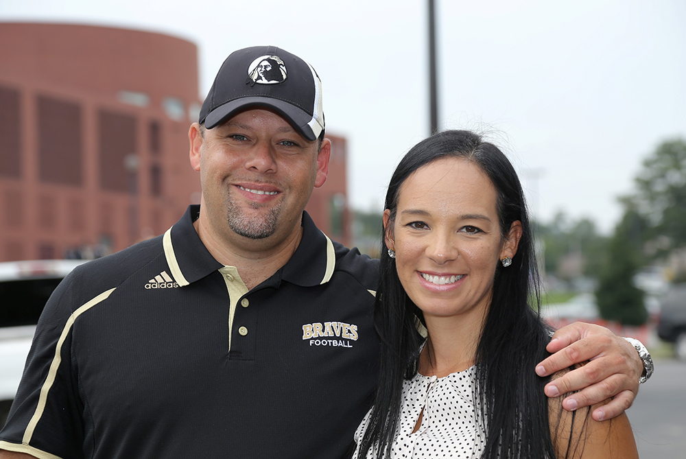 Two American Indian alumni in UNCP shirts. The man is wearing a Braves Football polo shirt and UNC Pembroke hat with his arm around the woman. They're smiling at the camera.
