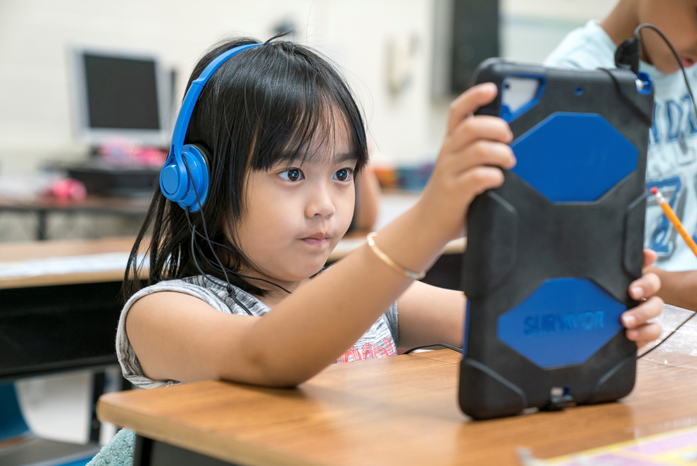 Young, elementary school student at her desk in the classroom, wearing headphones and holding a tablet.