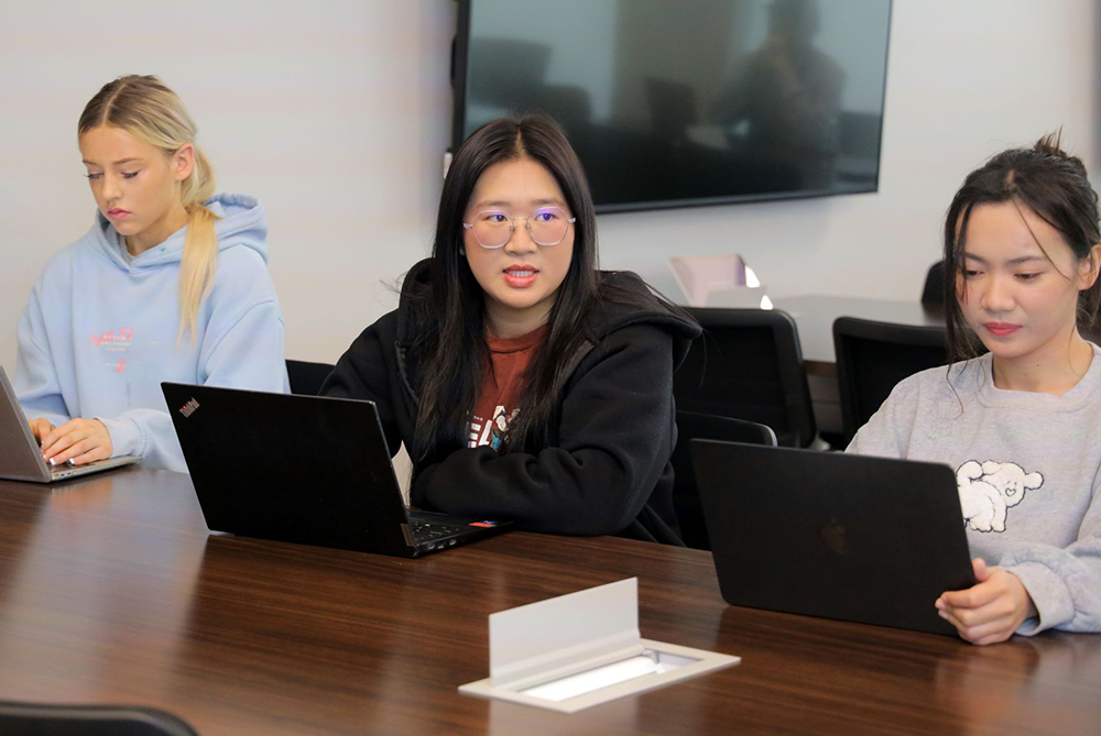 International students in class, seated at laptops. One student is speaking to the professor who is off-screen.