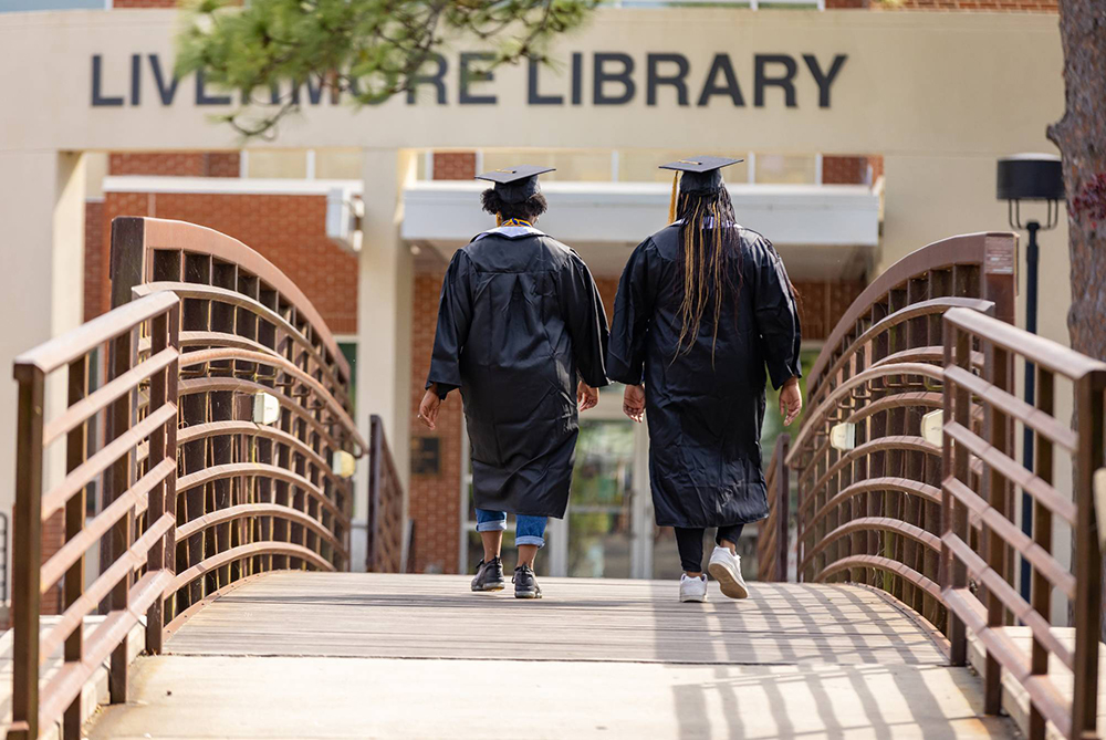two graduates in gowns walking across the UNCP bridge