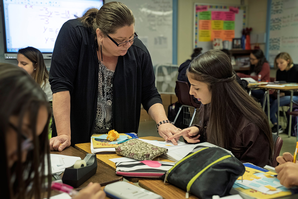 Middle School teacher leans over a group of desks to help a student with an assignment.