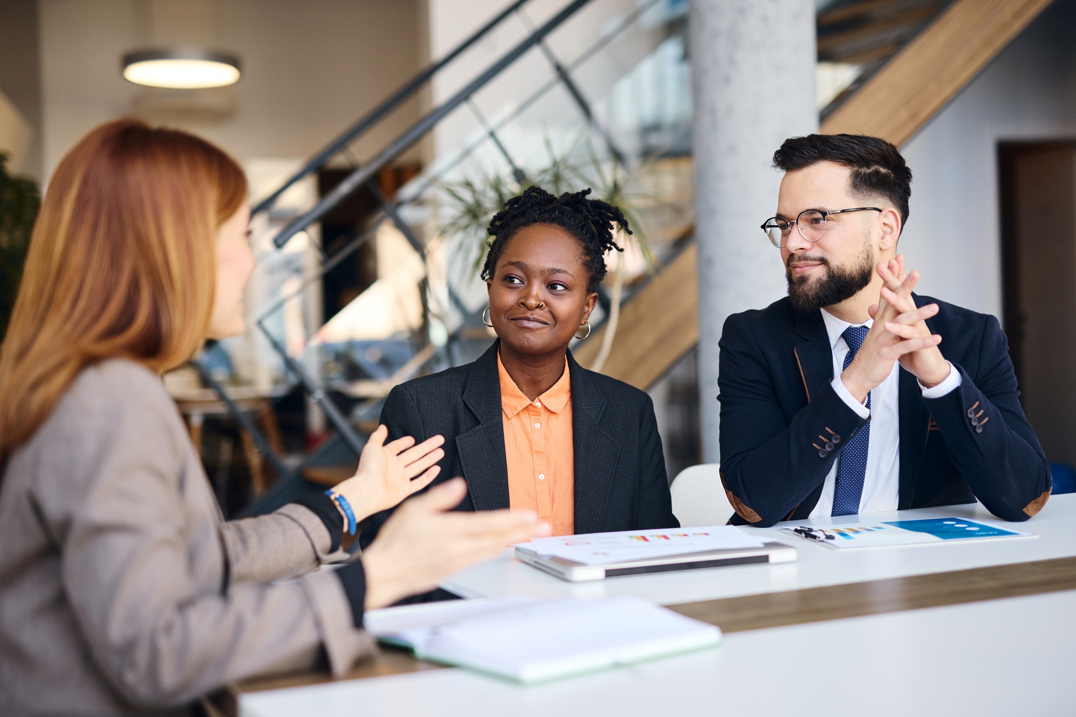Three people in business attire at a table in an office space. One long-haired woman sits to the left, facing a black woman and a white man. She's gesturing as she explains something.