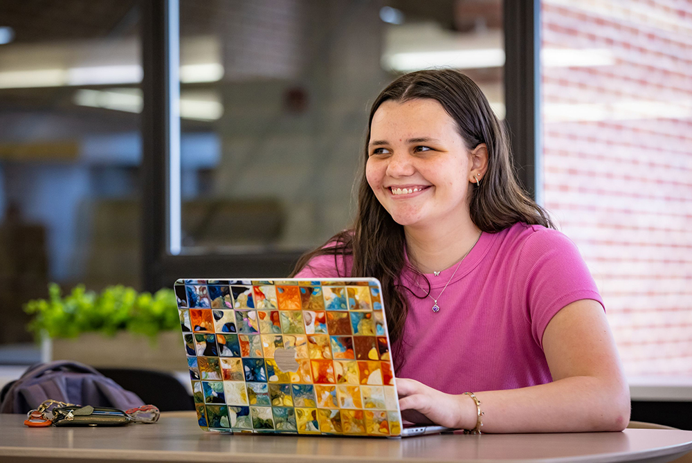 Female student at a table with a laptop open in front of her. Her laptop has a colorful case. She's smiling and looking to her right.