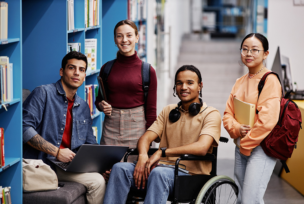 Students grathered in library. A male is seated on a bench with a laptop open. Two females are standing, wearing backpacks and holding study materials. A male is setad in a whellchair wearing headphones around his neck. All are facing the camera and smiling.