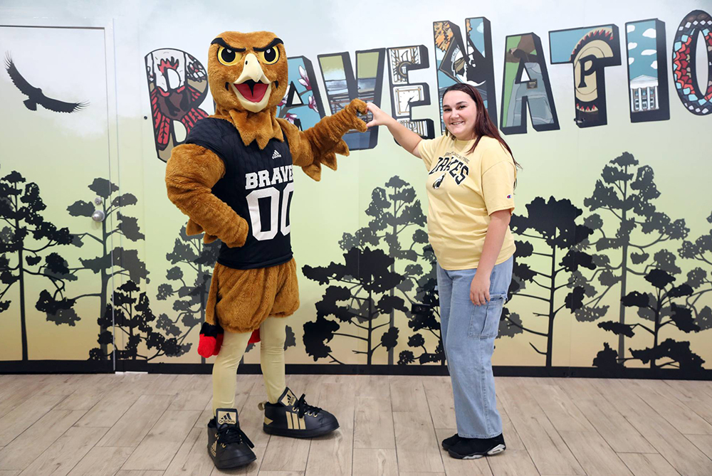 Bravehawk and a student stand in front of a postcard mural