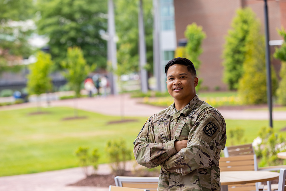 male soldier in uniform standing outdoors with greenery and outdoor furniture in the background.
