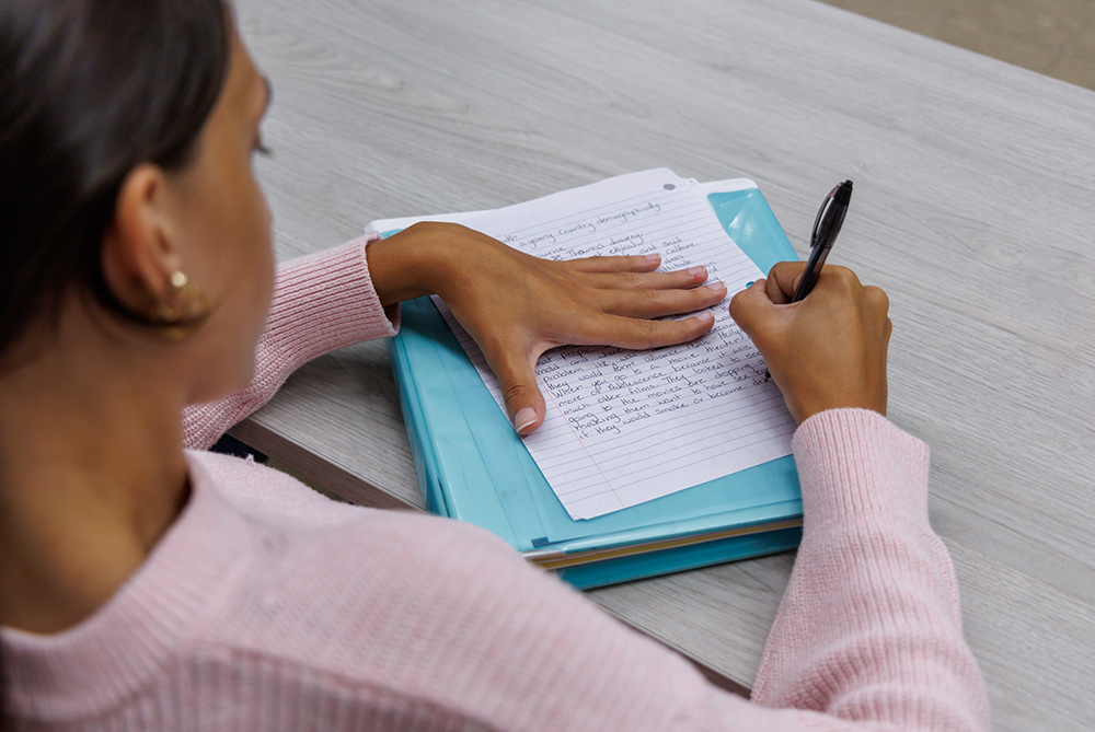 over the shoulder view of a female student writing in a notebook on a desk in a classroom
