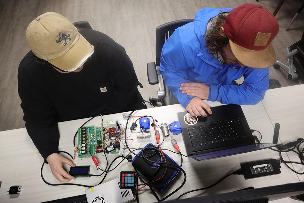 Overhead view of two male students in baseball caps seated at a table with a spread of electronics in front of them, working on a laptop.