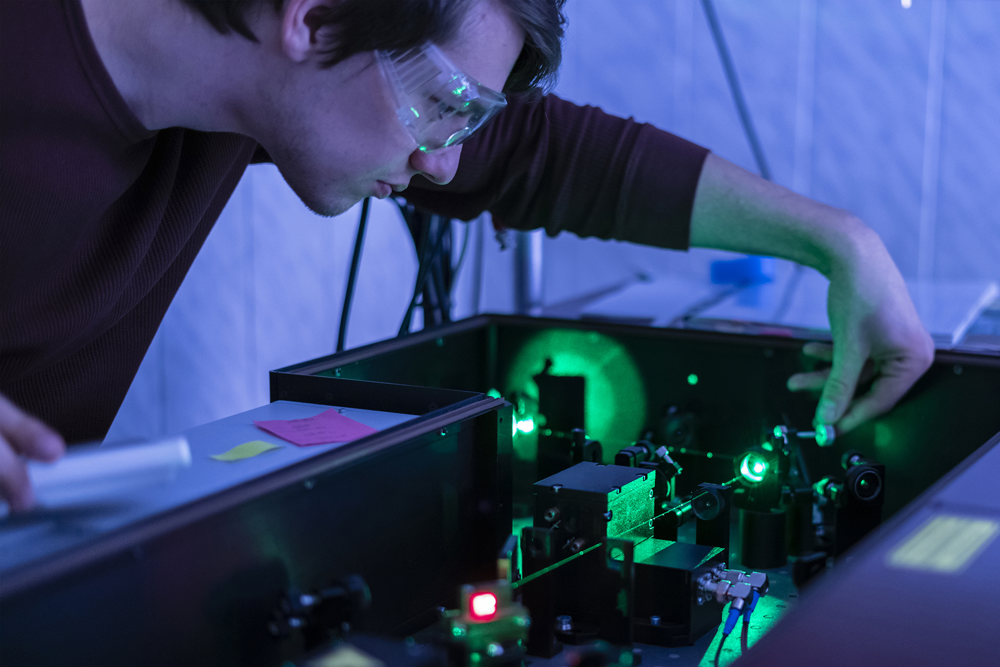 Male student wearing goggles leans over a green laser machine on the campus of UNCP.