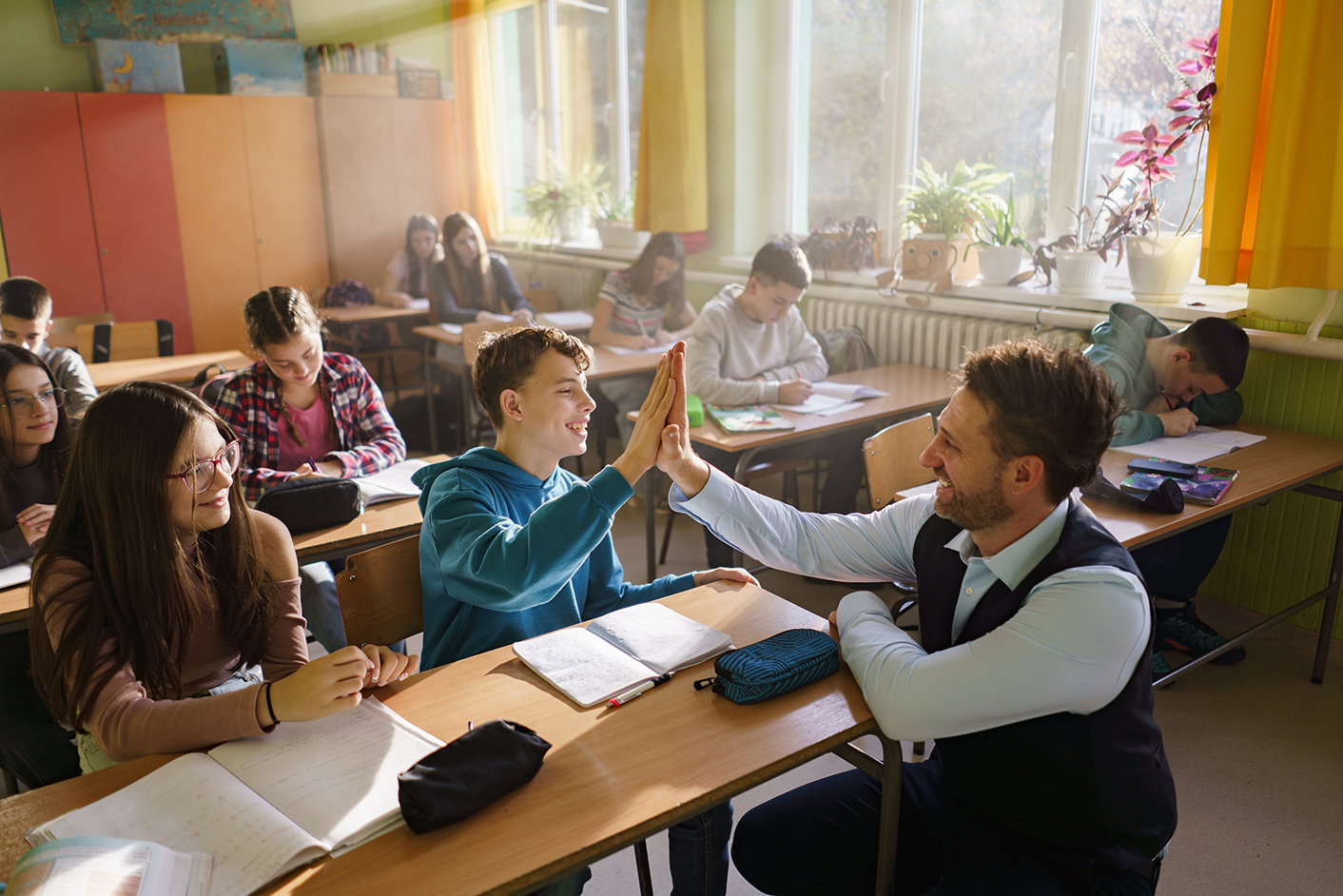 teacher in a classroom with students and high fiving a student while knealing at his desk