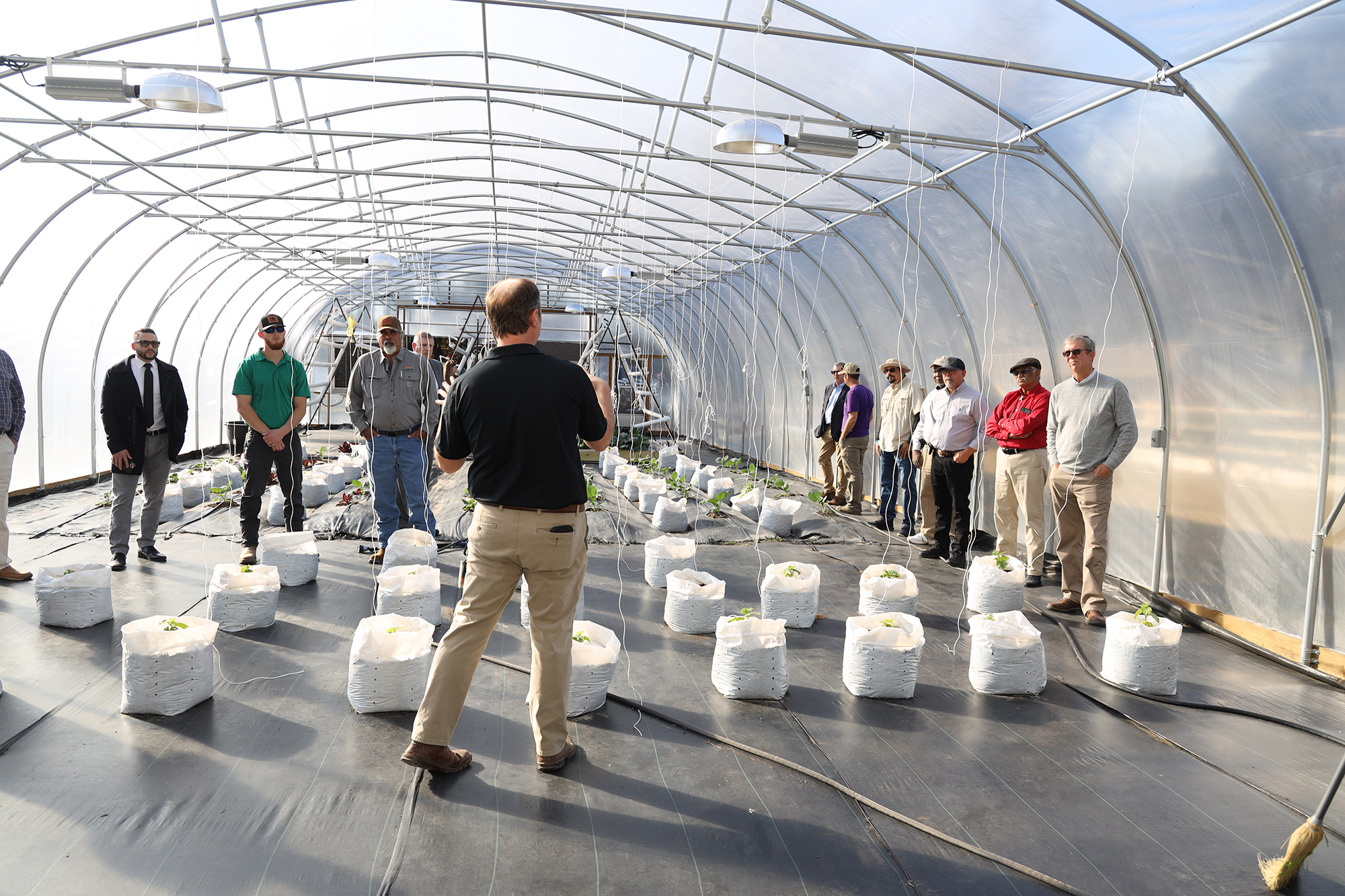 Dr. Bryan Sales addresses a group of visitors inside a UNCP agricultural greenhouse facility, with rows of potted plants lining the floor, representing the program's research and industry partnerships.