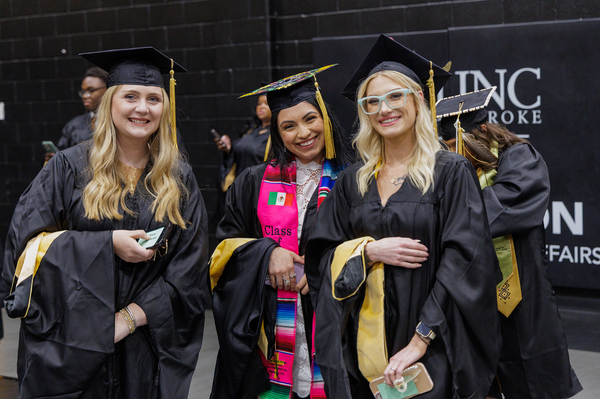 Three female students in graduation attire, smiling for the camera and holding their hoods.
